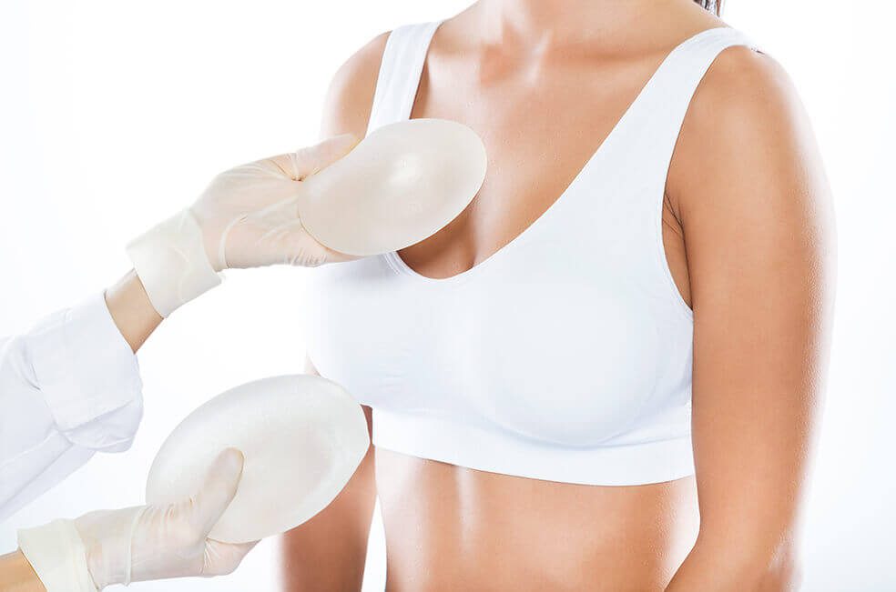 A woman in white bra holding up two breast implants.